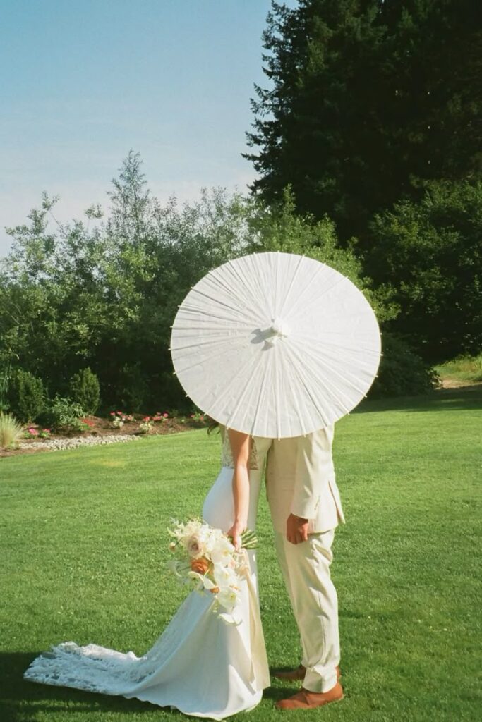 Bride and groom under a white parasol in a lush garden setting.
