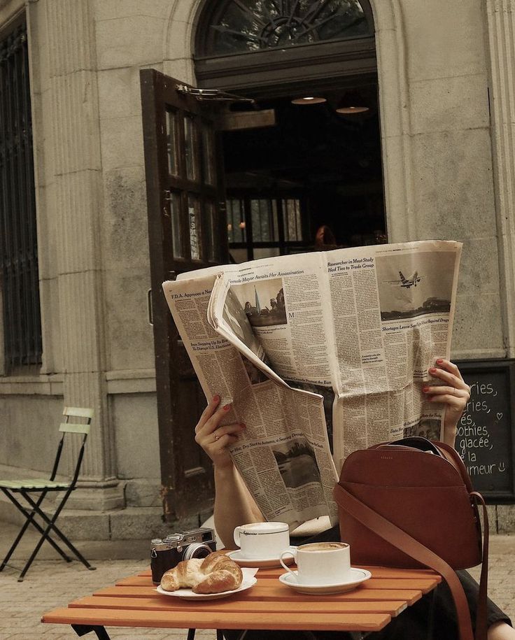 Person reading newspaper at a café table with coffee, croissants, and a vintage camera outdoors.
