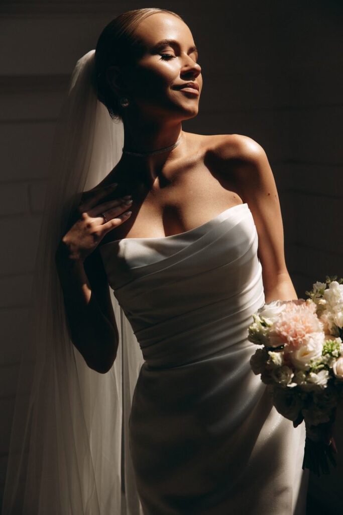 Bride in elegant white dress holding bouquet, illuminated by warm light, radiating joy and elegance on her wedding day.