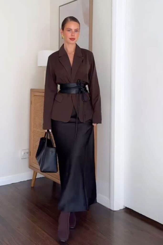Woman in stylish brown blazer and long black skirt, accessorized with a belt and handbag, standing in a chic room.
