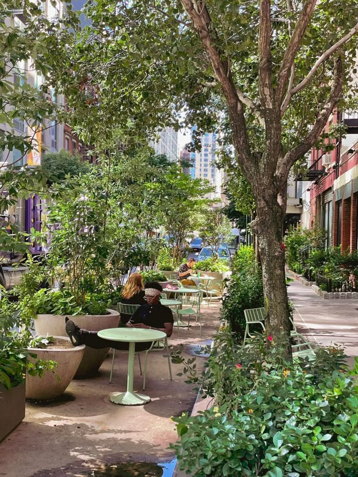Urban outdoor café surrounded by lush greenery and trees, with people relaxing at tables in a vibrant city setting.