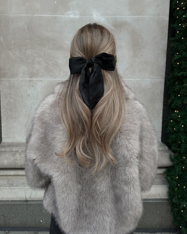 _ 13 Woman with long hair tied with a black bow wearing a gray fur coat, viewed from the back, against a stone wall. | Sky Rye Design Woman with long hair tied with a black bow wearing a gray fur coat, viewed from the back, against a stone wall.