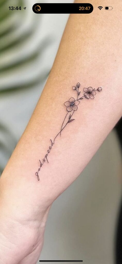 Delicate floral tattoo with godspeed script on forearm. Minimalist black ink design.