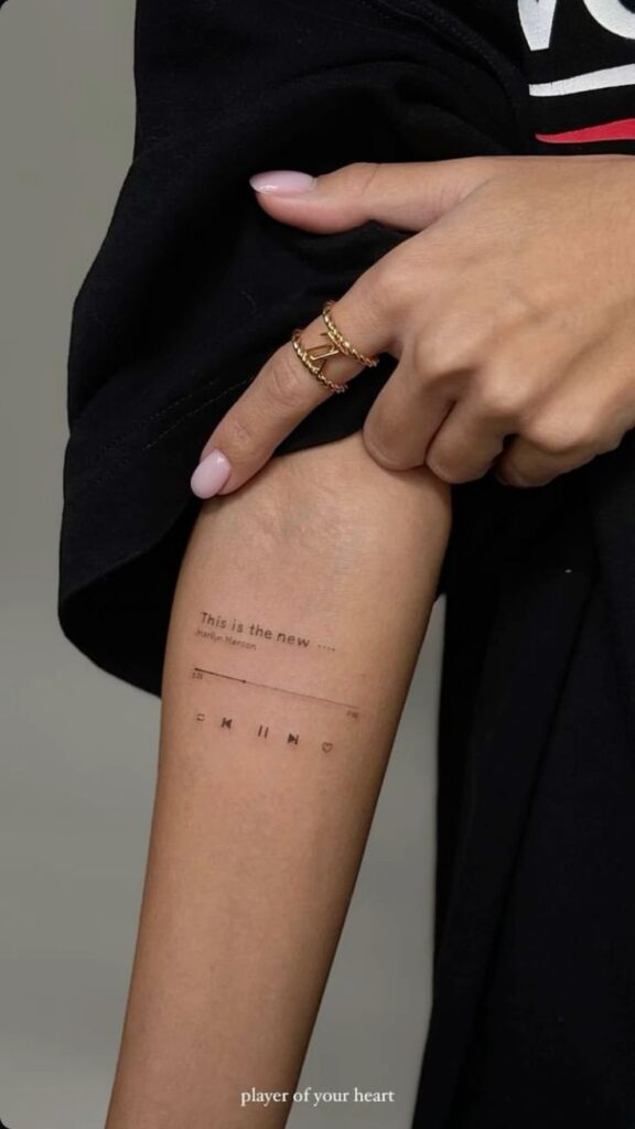 Arm tattoo resembling a music player, with text and icons on skin. Minimalistic design highlighted by a golden ring.