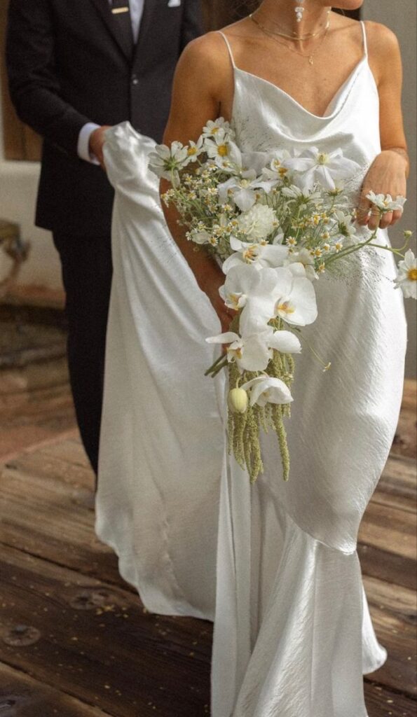 Bride in elegant white dress holds flower bouquet, groom in suit assists, on rustic wooden floor.