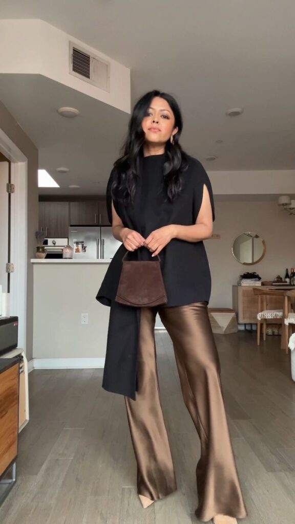 Woman in stylish black top and brown satin pants holding a bag, standing in a modern living room.
