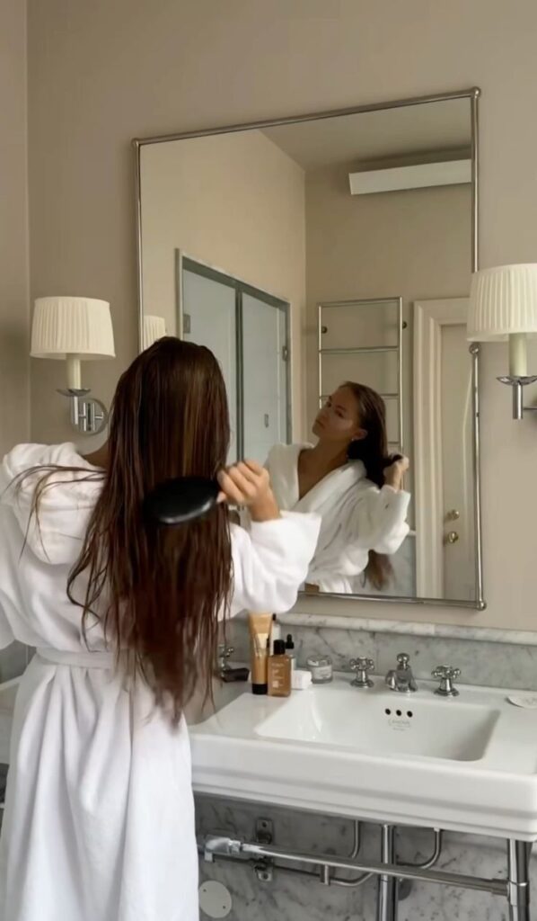 _ 14 Woman in white robe brushing long hair in front of bathroom mirror, with skincare products on marble sink. | Sky Rye Design Woman in white robe brushing long hair in front of bathroom mirror, with skincare products on marble sink.