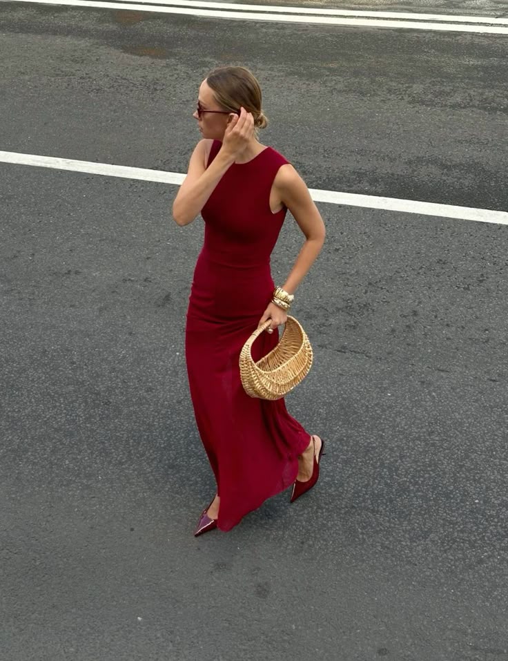 Woman in a red dress with wicker handbag walking on street, wearing sunglasses and high heels.
