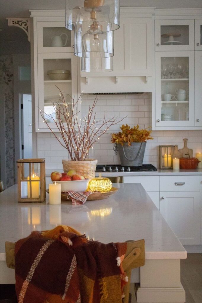 Cozy kitchen with autumn decor: candles, apples, and lights on a white counter, accented by warm fall colors and textures.