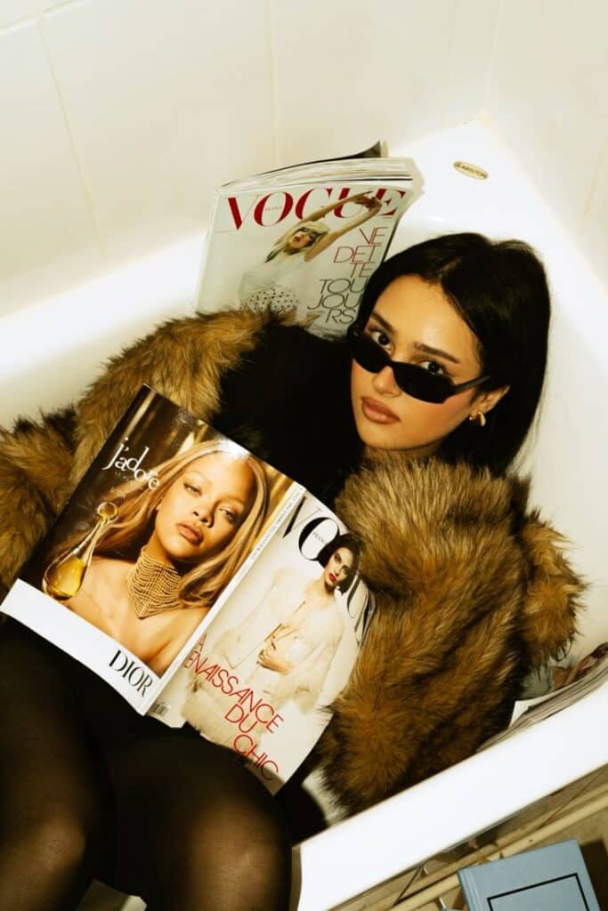 Woman in sunglasses and fur coat reading Vogue magazine in a bathtub.