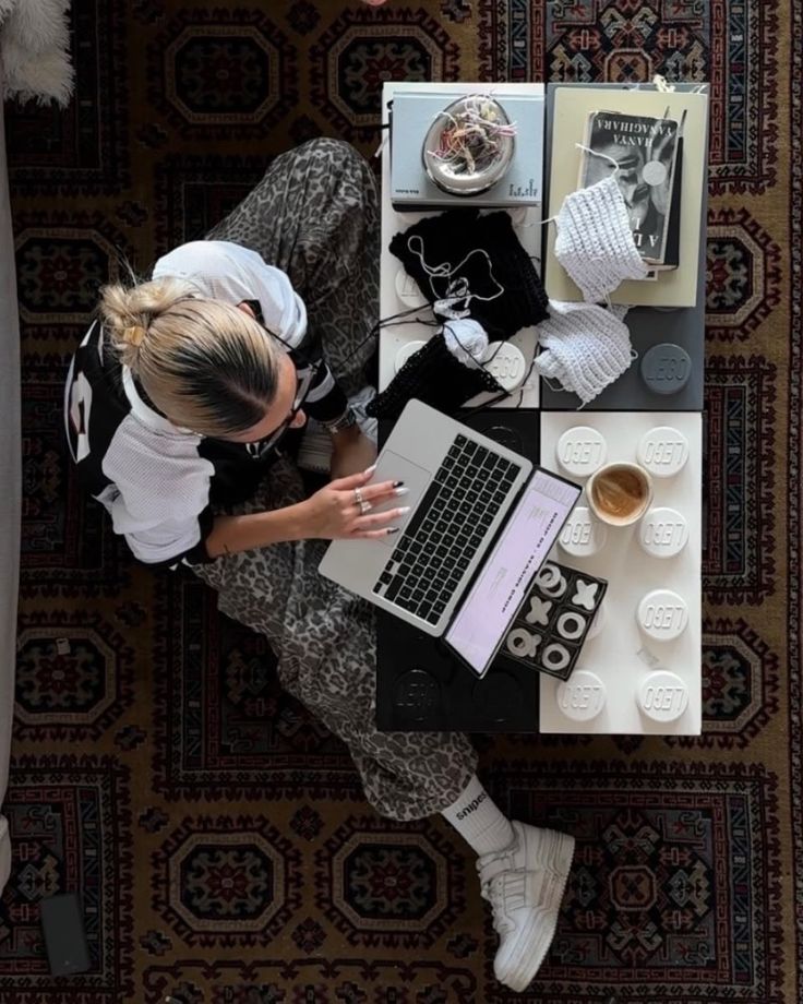 Person using a laptop on a coffee table, surrounded by books, knitting materials, and a coffee cup in a cozy setting.
