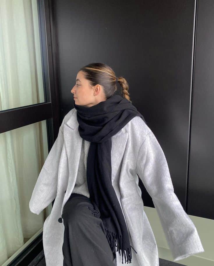 Woman in a gray coat and black scarf sits in front of a window, looking to the left.