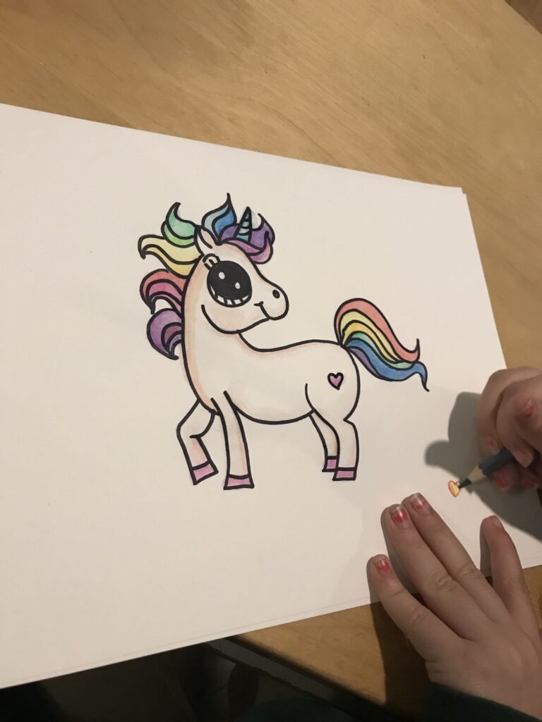 Child coloring cheerful unicorn with rainbow mane on white paper.
