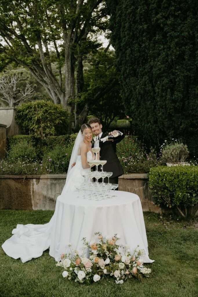 Bride and groom pouring champagne tower at outdoor wedding, surrounded by lush greenery.