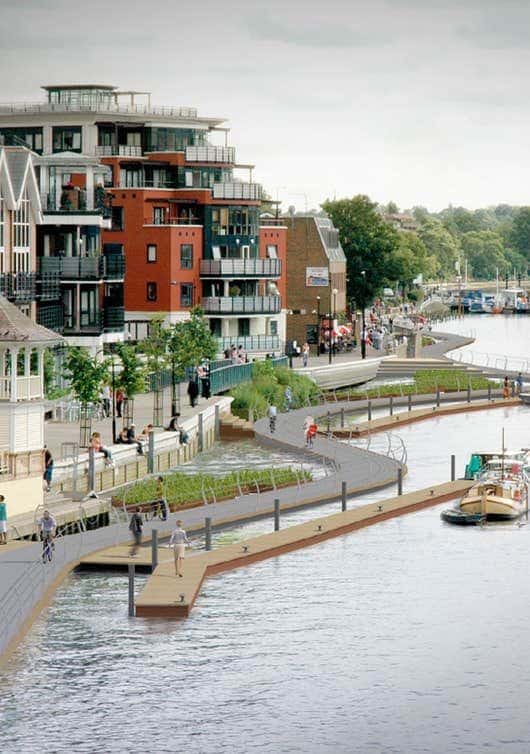 Waterfront promenade with modern apartments and walking path, featuring boats and greenery under a cloudy sky.