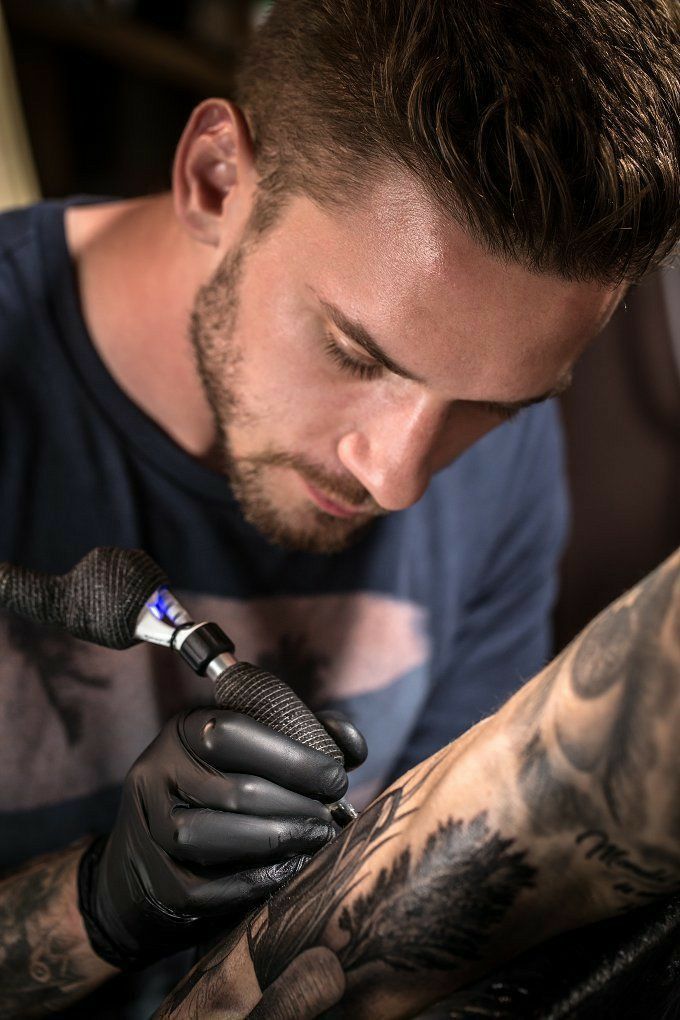 Tattoo artist focused on creating intricate arm design, wearing gloves, using tattoo machine.