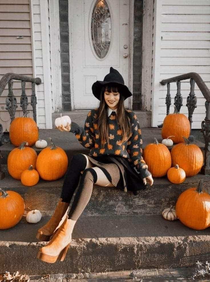 Woman in witch hat sitting with pumpkins on porch steps, autumn fashion, cozy fall vibes.