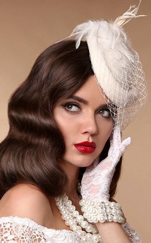 Vintage elegance: Woman in lace gloves, pearl jewelry, red lipstick, and feathered hat against beige background.