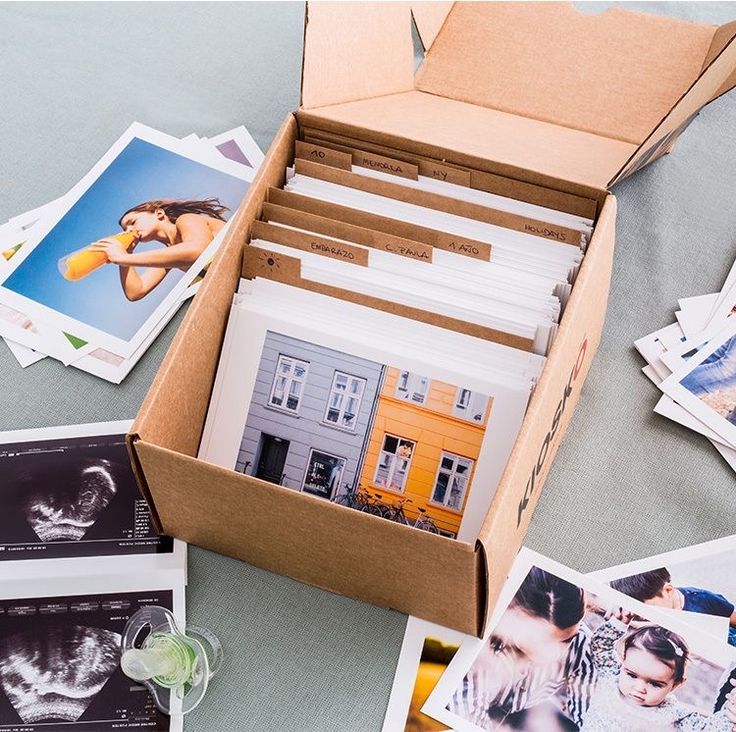 Box of printed photos and ultrasound images on a table, including a woman drinking orange juice and colorful buildings.