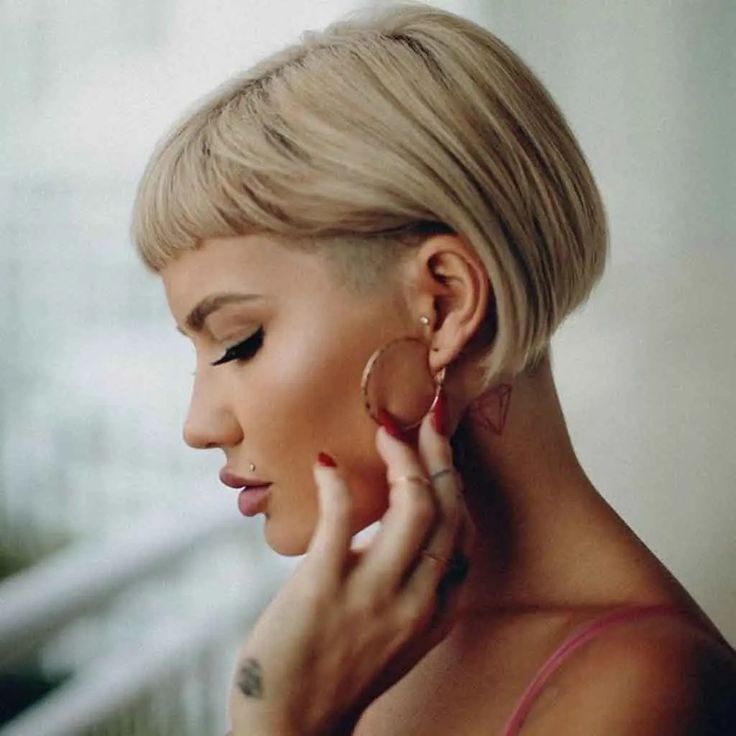 Woman with short blond bob hairstyle, holding a hoop earring, showcasing edgy piercings and makeup.