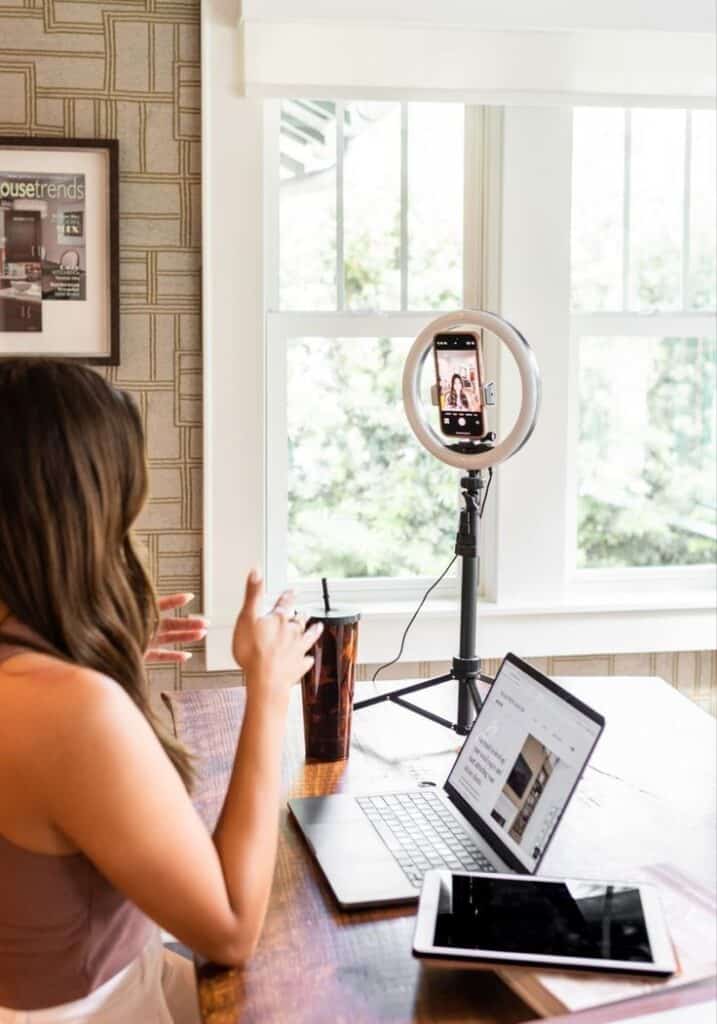 Woman video conferencing from home with laptop, tablet, and ring light setup near window.