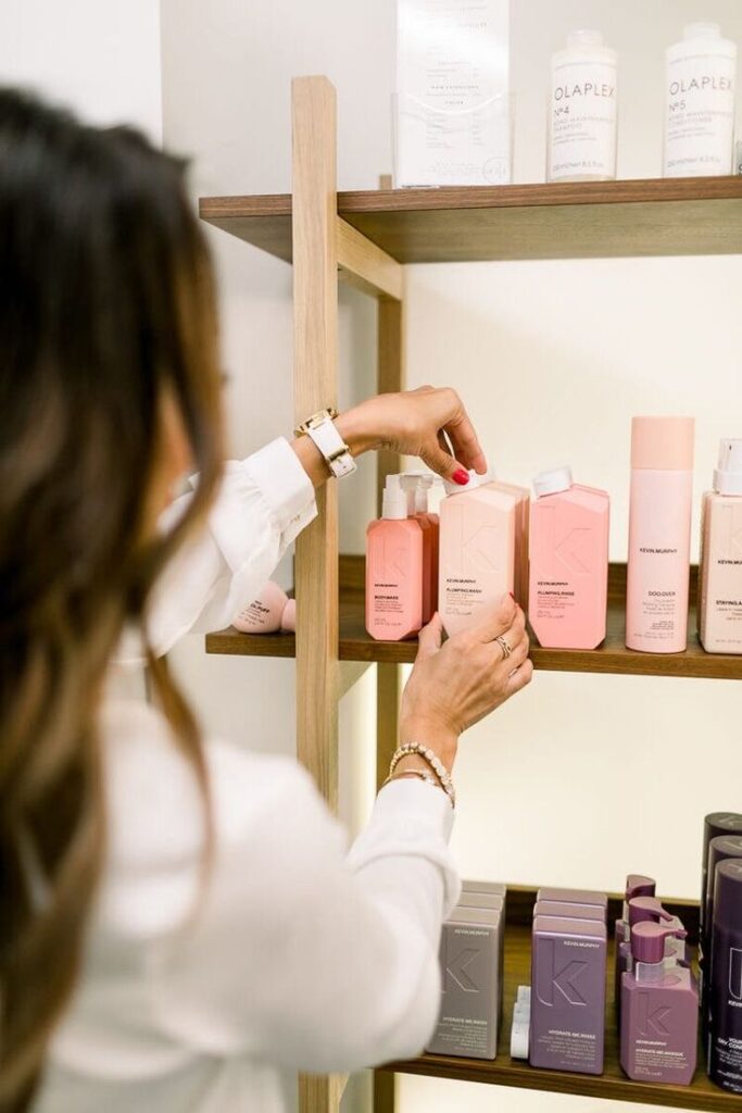 _ 16 Person organizing haircare products on a wooden shelf, focusing on colorful shampoo bottles. | Sky Rye Design Person organizing haircare products on a wooden shelf, focusing on colorful shampoo bottles.
