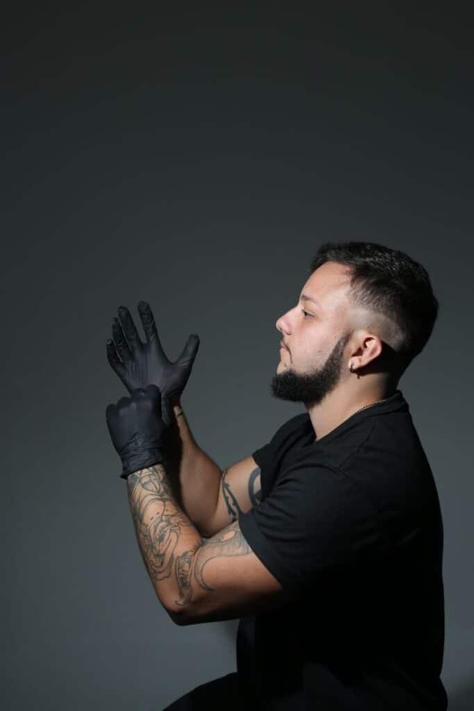 Man with tattoos wearing black gloves, ready for work against a dark background.