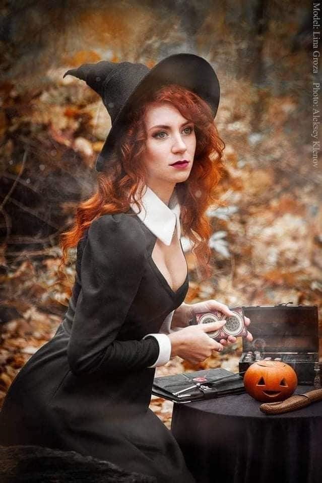 Witch in black dress with red hair holding a pocket watch in autumn forest, Jack-o'-lantern on table nearby.