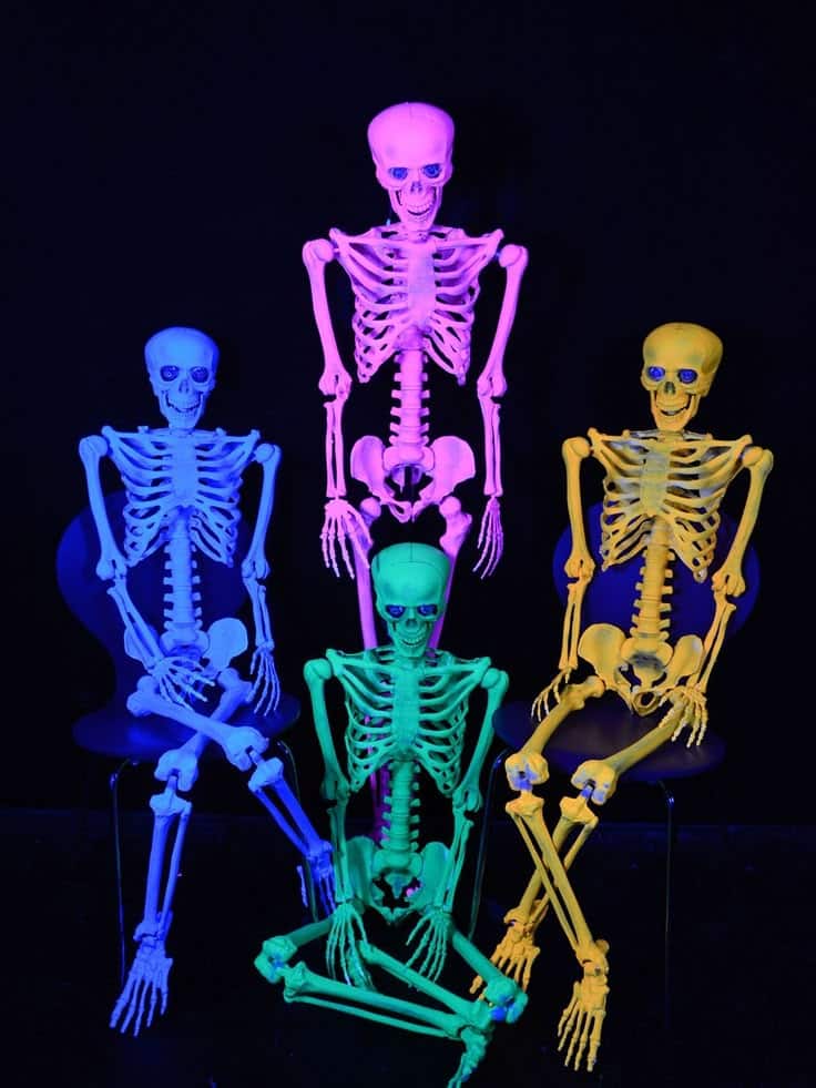 Colorful neon skeletons under blacklight, sitting and posing against a black background, create a vibrant Halloween scene.