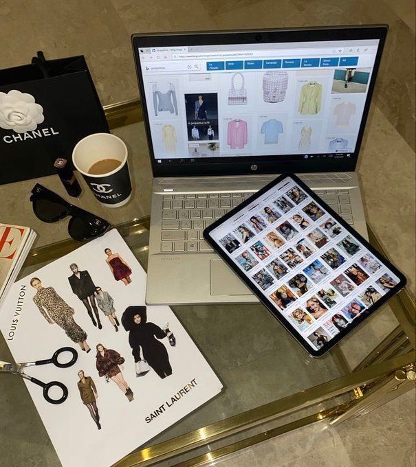 Fashion workspace with laptop, tablet, Chanel coffee cup, and designer lookbook on a glass table.