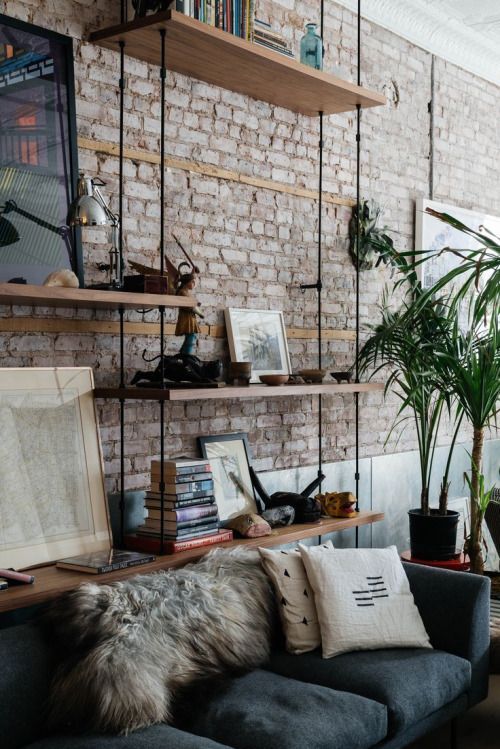 Cozy living room with brick wall, floating shelves, books, plants, and a gray sofa adorned with pillows and a fur throw.