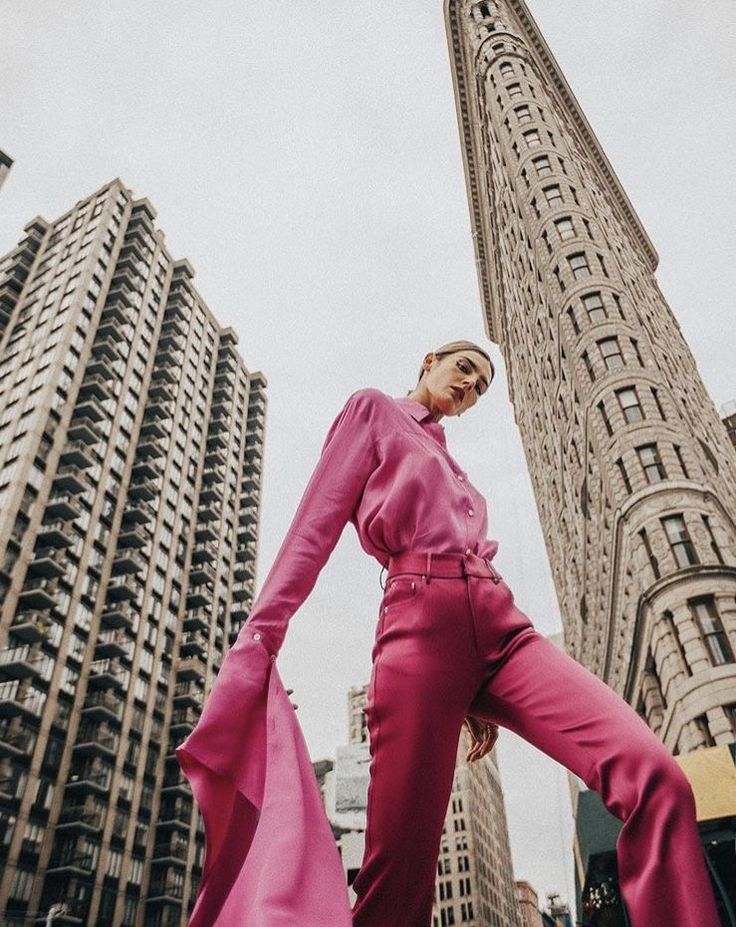 Fashionable figure in vibrant pink outfit stands confidently in city with towering historic buildings in the background.