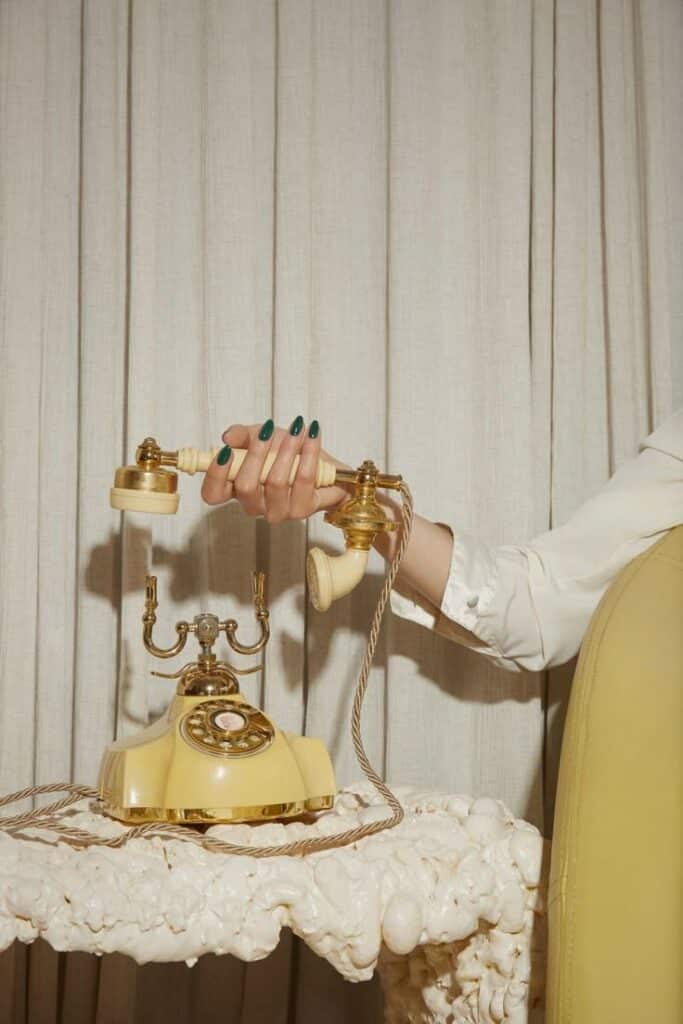 Vintage yellow rotary phone on ornate table with hand holding receiver, elegant green nail polish.