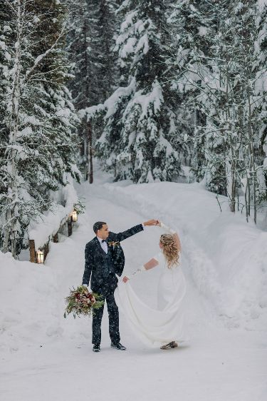 Couple dances in snowy forest on their wedding day, surrounded by snow-covered trees, holding a bouquet.