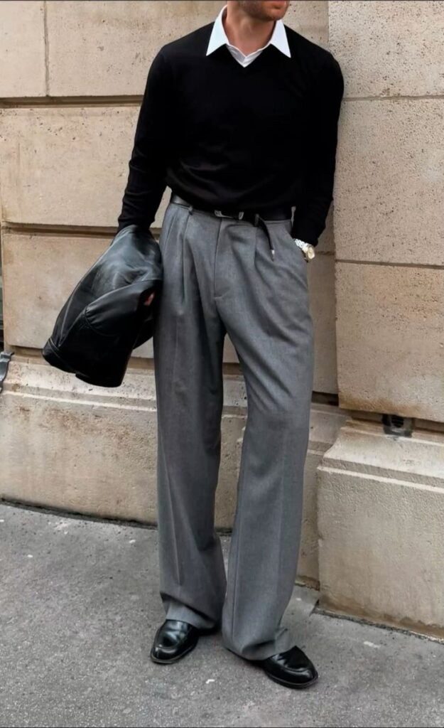 Stylish man in black sweater and gray pants holding a leather jacket, leaning against a stone wall.