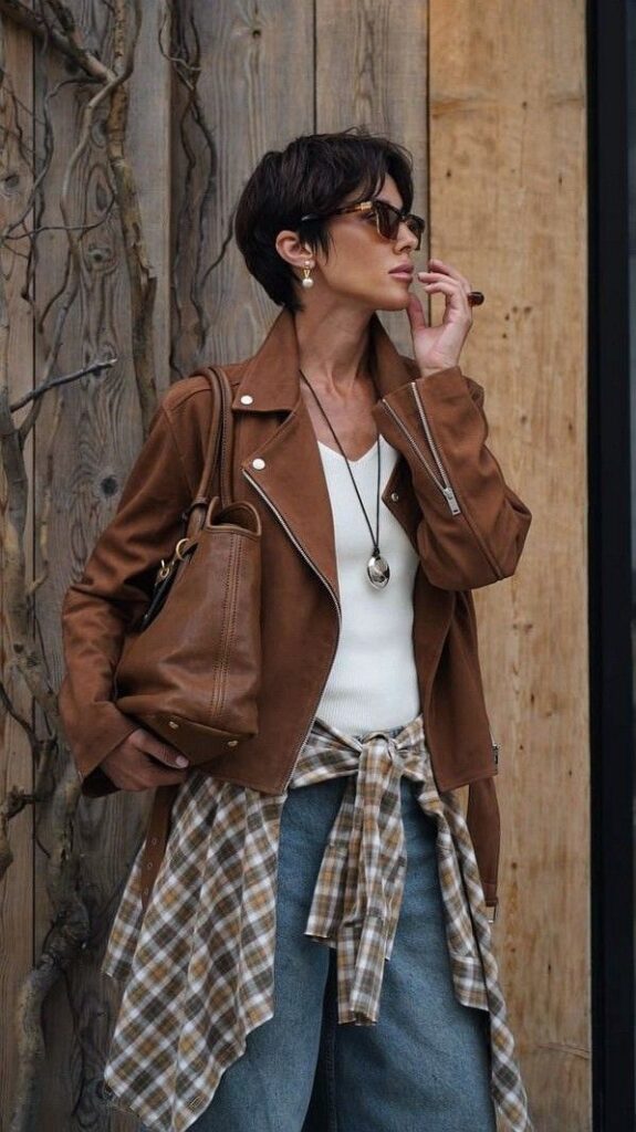 Fashionable woman in brown jacket and sunglasses, wearing plaid shirt and denim, standing against wooden background.