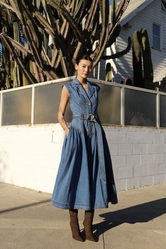 Woman in denim dress and boots stands outdoor near tall cacti, with a modern house backdrop, under clear blue sky.
