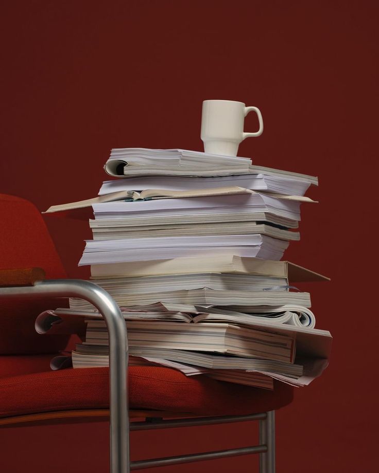 Stack of books and papers with a white mug on a red chair against a maroon background.