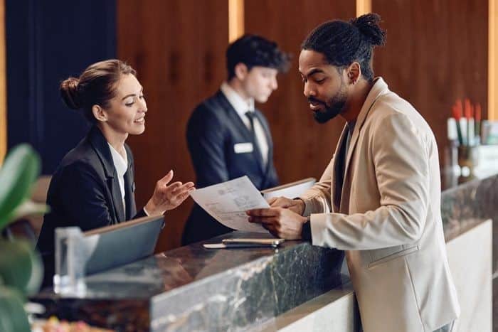 Man at hotel reception desk checking in, assisted by a receptionist while another staff member works in the background.