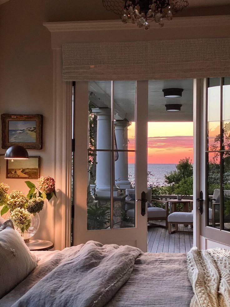 17d7c12c-d9ae-487b-ac51-eb309f19422a Cozy bedroom with open doors to a stunning sunset view over the ocean, featuring flowers and inviting decor. | Sky Rye Design Cozy bedroom with open doors to a stunning sunset view over the ocean, featuring flowers and inviting decor.