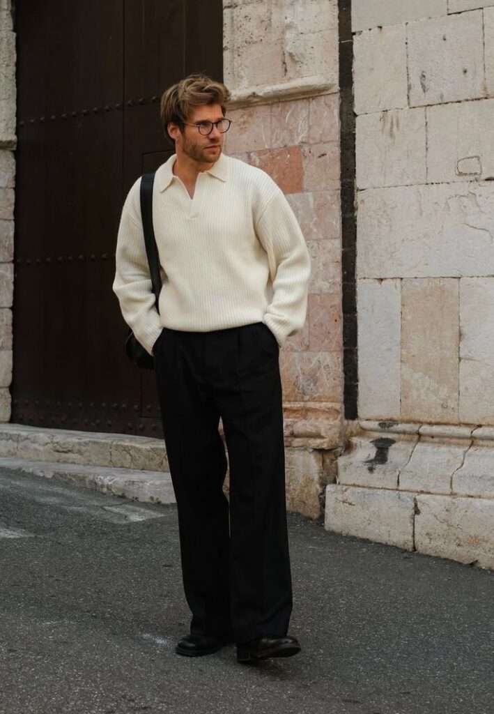 Man wearing a cream sweater and black trousers standing on a street near a stone building.