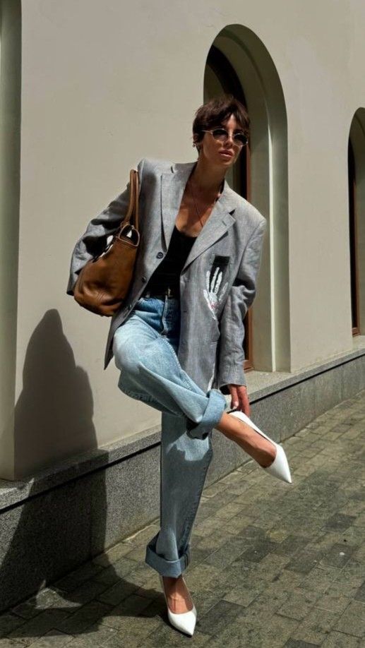 Stylish person in a gray blazer, jeans, and white heels posing outdoors with a brown bag and sunglasses.