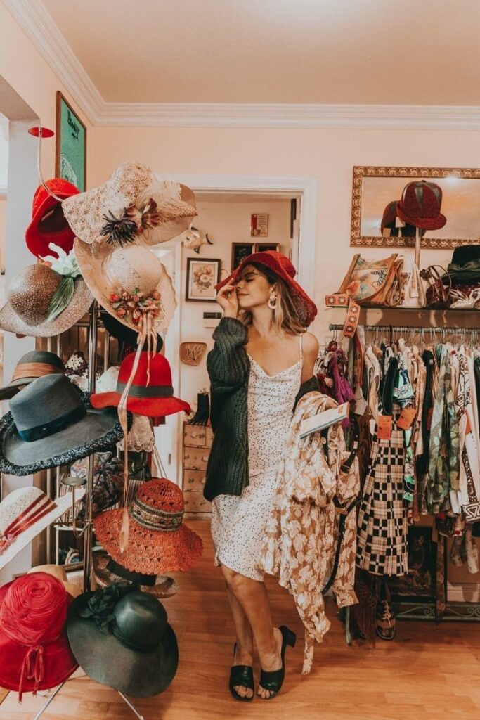 Woman browsing hats in a vintage store, wearing a red hat and floral dress, surrounded by colorful headwear.