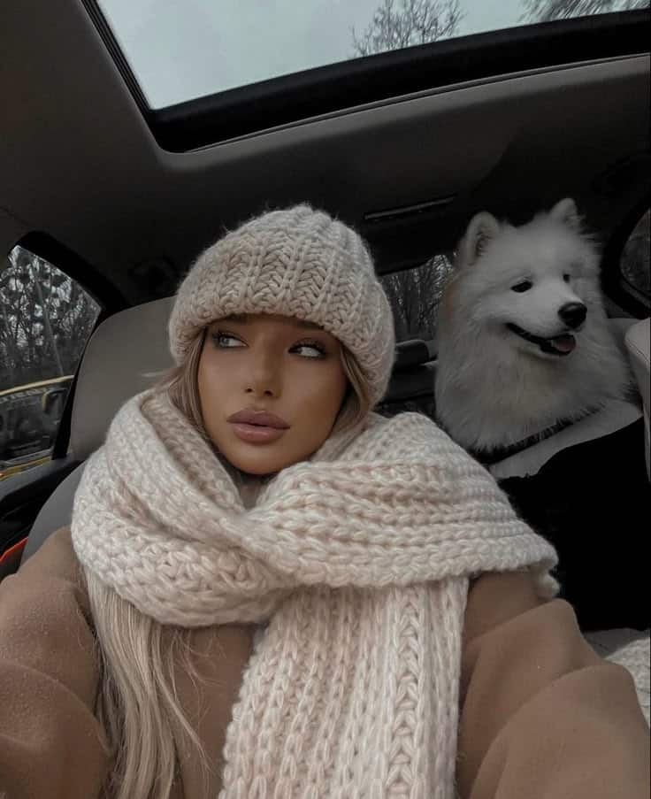 Woman in cozy knitwear and hat sitting in a car with a fluffy white dog on a winter day.