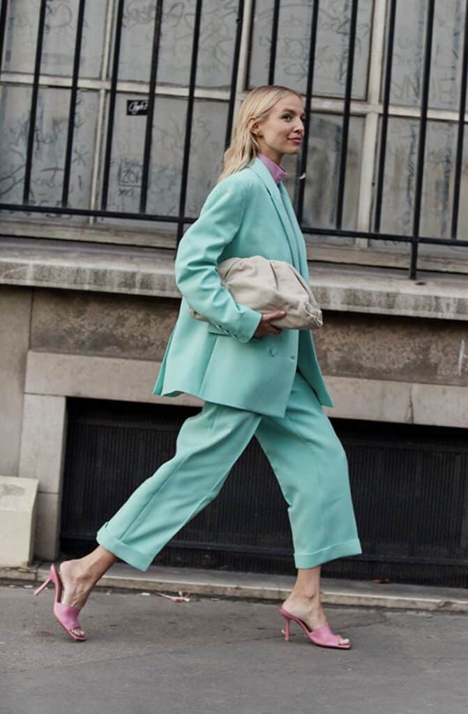 Woman in turquoise suit and pink heels walking confidently on city sidewalk, holding a beige handbag. Fashion street style.