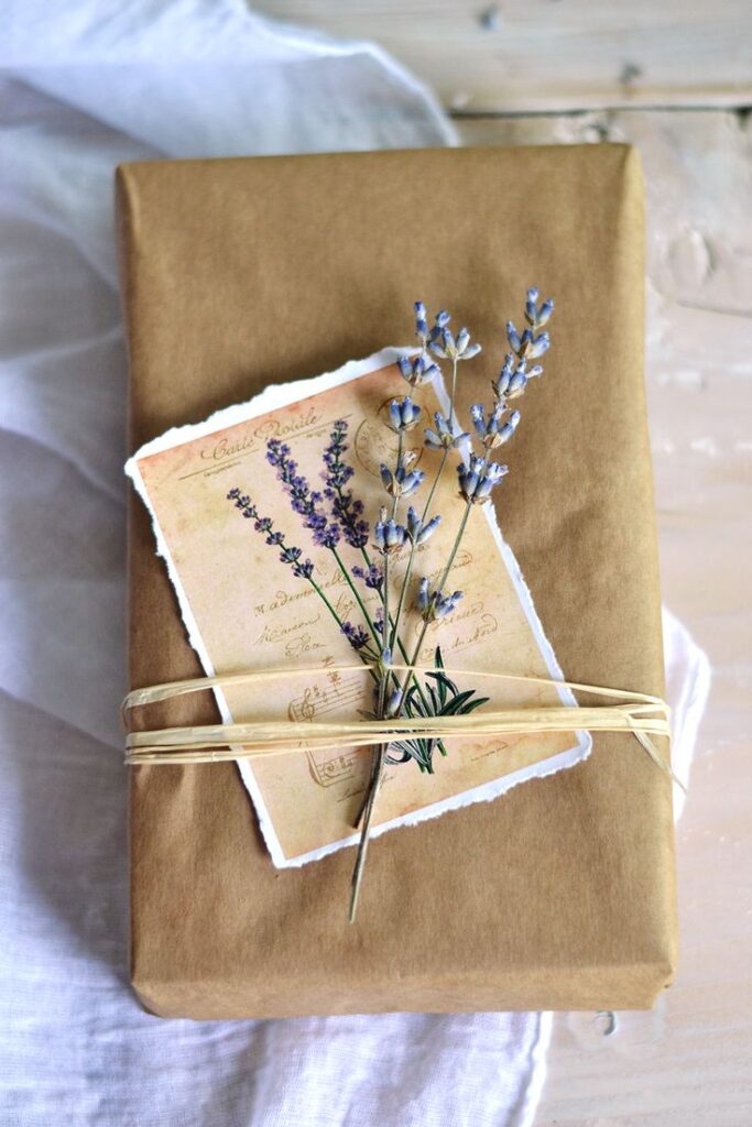 Brown paper gift with lavender sprigs and vintage card, tied with raffia on white fabric background.
