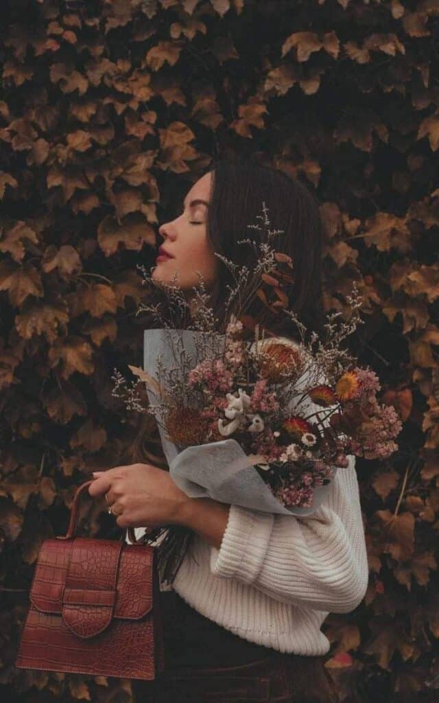 Woman in white sweater holding autumn flowers and a brown handbag, standing against a leafy background.