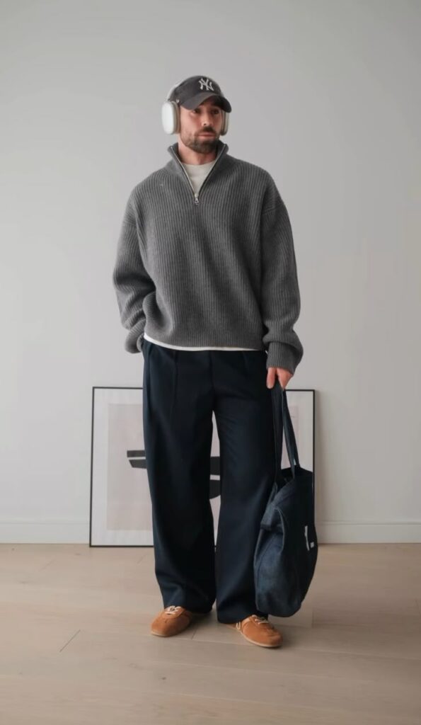 Man in casual fashion: gray sweater, baggy pants, headphones, cap, brown shoes, and tote bag in minimalist room.