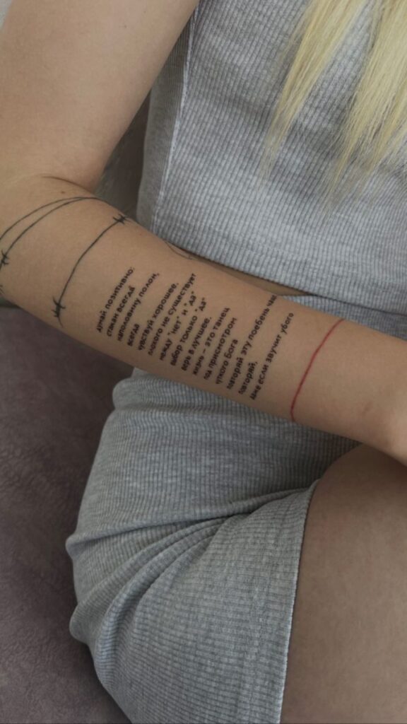 Person with tattooed text and barbed wire design on forearm, wearing a gray dress, sitting on a textured surface.