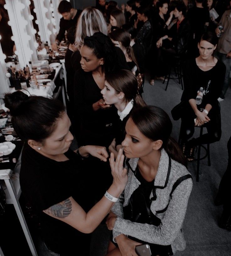 Backstage makeup session at fashion show with models and artists preparing, surrounded by beauty products.